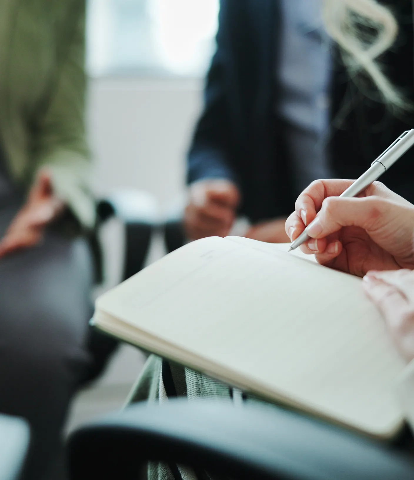Notizbuch in das gerade etwas notiert wird. Im Hintergrund noch zwei weitere Personen.