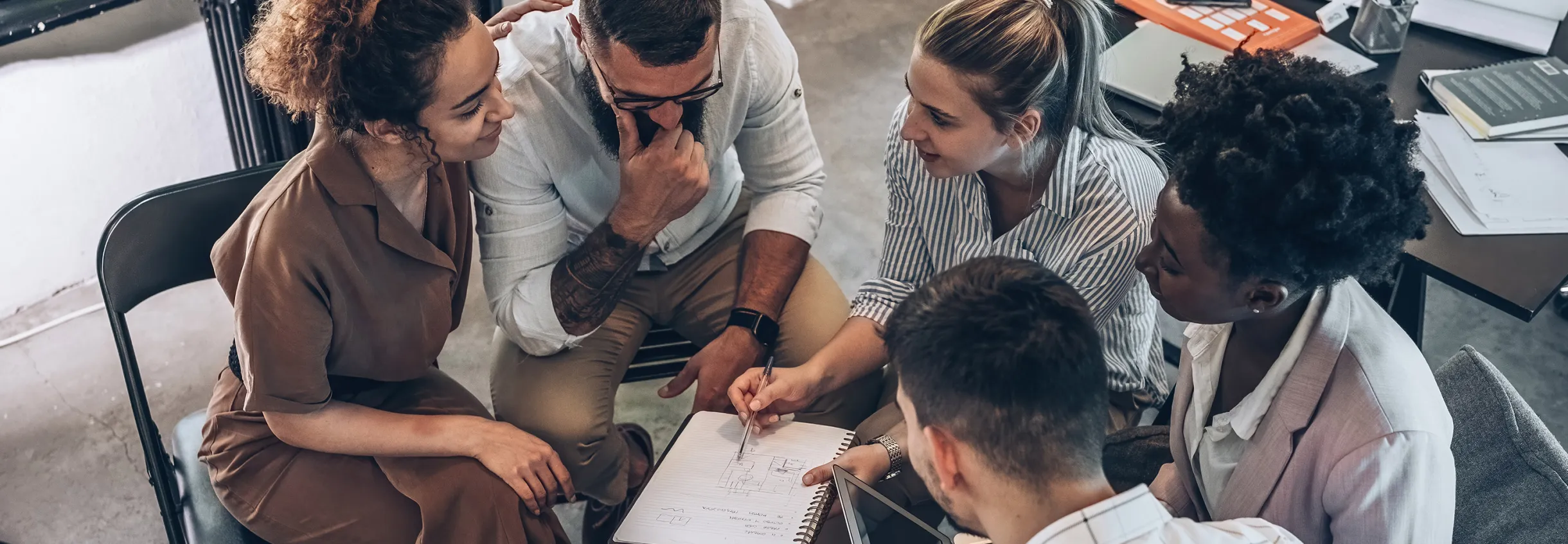Gruppe von f&uuml;nf Personen sitzt im Kreis und diskutiert und halten ein Notizbuch in der Hand.